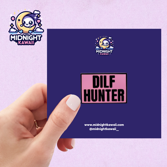 A pink rectangular shaped enamel pin with the words "DILF HUNTER" engraved in black. Size 3-4cm. The pin has a securing arrangement on the back to enable attachment to clothes, bags etc.