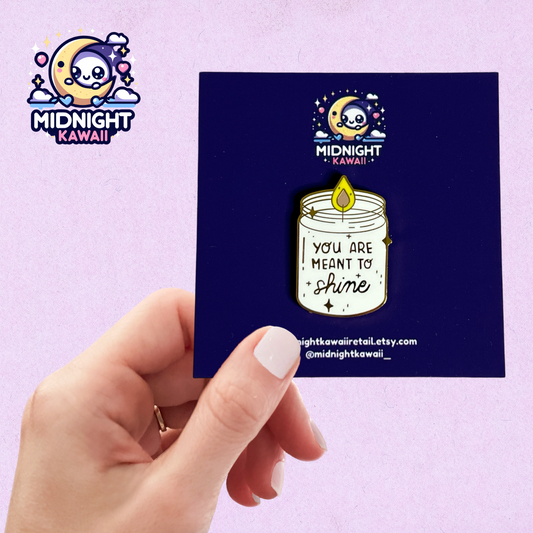 A white candle enamel pin with metallic gold text and pin outline. The candle is aflame with a yellow flame. The words "you are meant to shine" are engraved on the candle. Size 3-4cm. The pin has a securing arrangement on the back to enable attachment to clothes, bags etc.